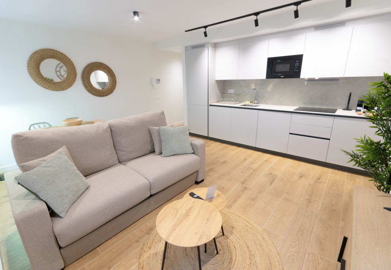 Salón con sofá, cojines verdes y cocina abierta al fondo, apartamento Urban Plus